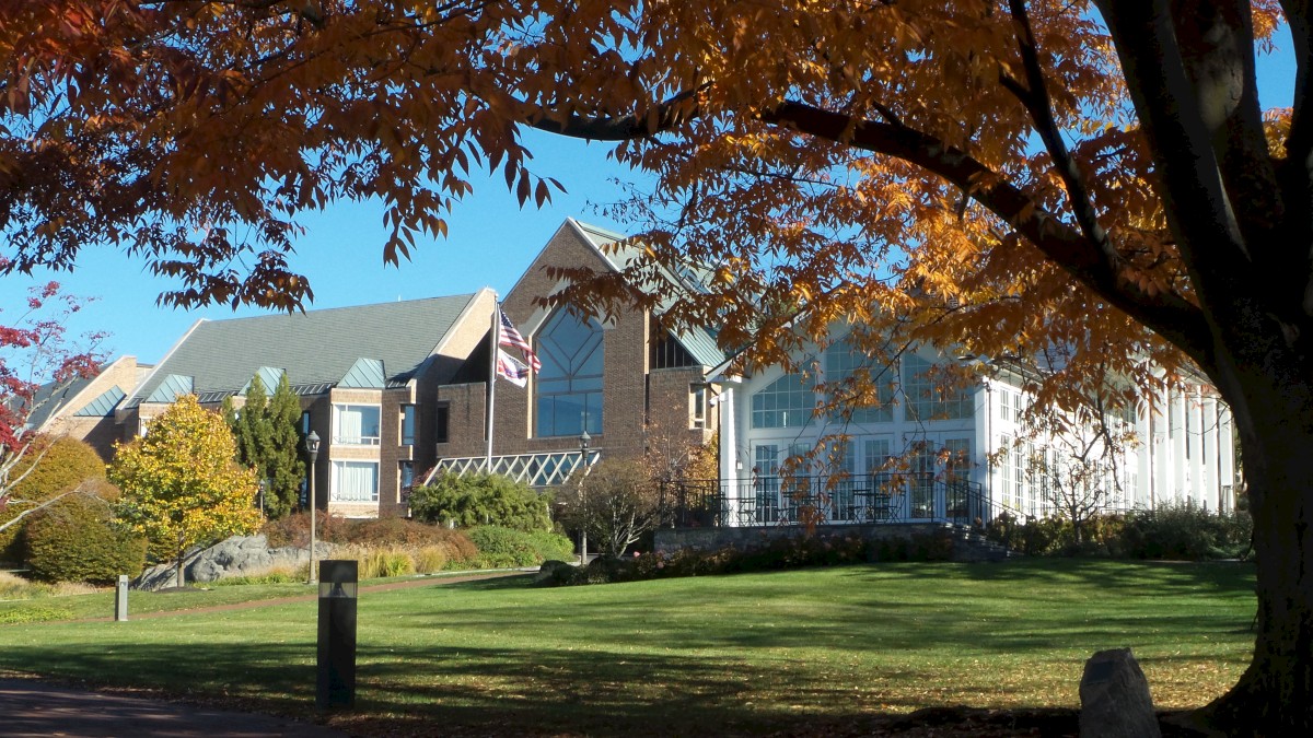 The image shows a campus building with fall foliage, a grassy lawn in the foreground, and a pathway leading towards the structure.